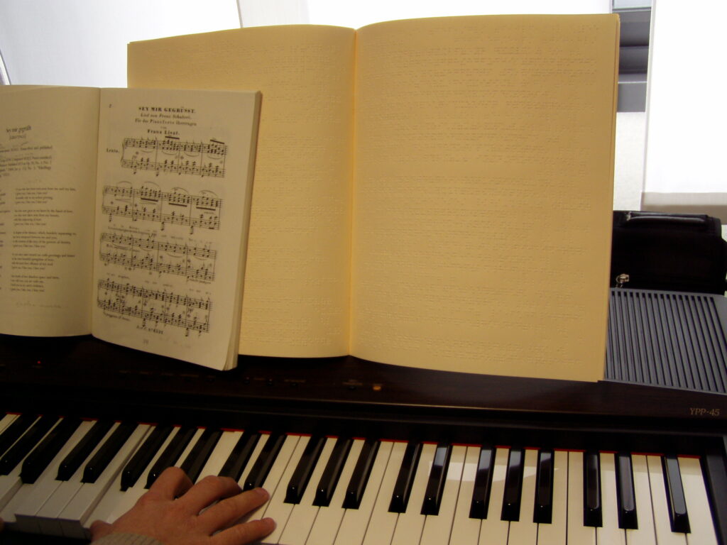 L'immagine raffigura un testo braille in nero e un testo di musica braille appoggiati ad una pianola